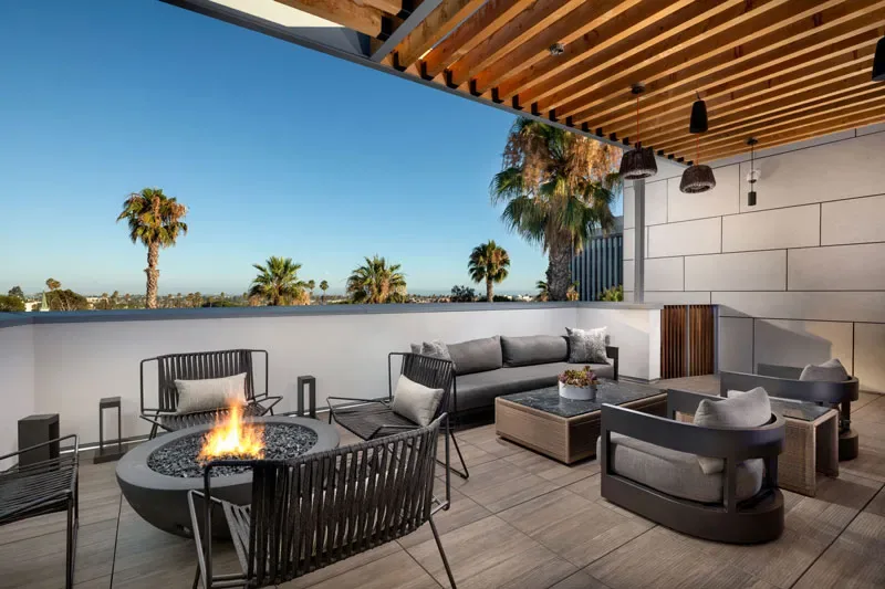 Rooftop Lounge With Fire Pit Seating, Cushioned Sofas, Wood Ceiling Beams, And Palm Tree Views.