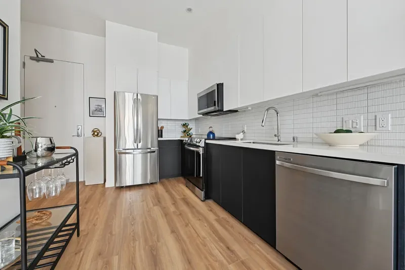 Modern Kitchen With Stainless Steel Appliances, White Tile Backsplash, Black Lower Cabinets, And Wood Flooring.